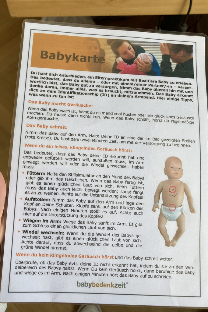 Laminierte Karte mit dem Titel "Babykarte". Sie enthält Pflegehinweise für ein RealCare Baby, inklusive Tipps zu Geräuschen, Bedürfnissen und ID-Erkennung.