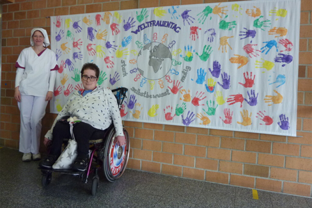 Die beiden Frauenbeauftragten präsentieren stolz ihr gemeinschaftlich gestaltetes Kunstwerk: eine Leinwand voller farbiger Handabdrücke, die hinter ihnen sichtbar ist.