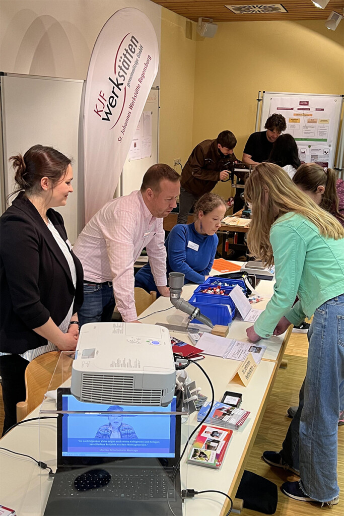 Die Mitarbeiter*innen zeigen zwei Besuchern am Messestand die Arbeit der Werkstätte.