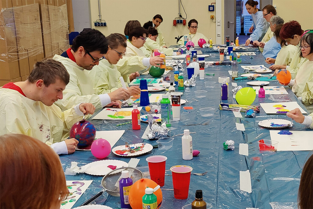 An einem langen Tisch verzieren viele Mitarbeiterinnen und Mitarbeiter Luftballons mit Pinsel und Farbe..