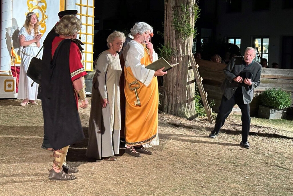 Eine Szene aus dem Theaterstück Der Brandner Kaspar und das ewig Leben. Ein Schauspieler liest aus einem goldenen Buch vor. Einige andere hören gespannt zu.