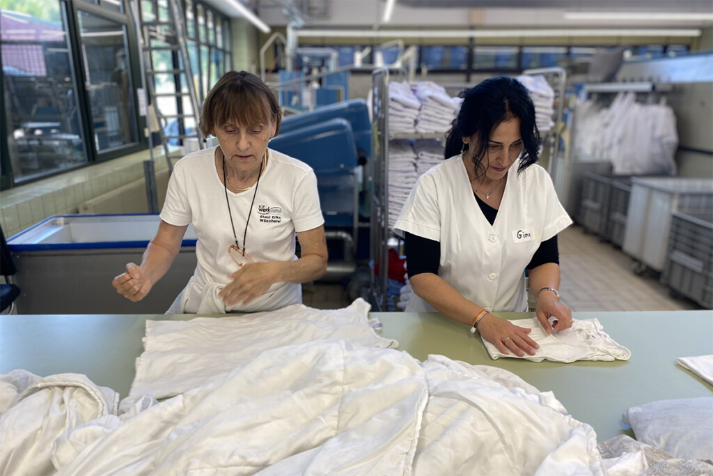 Feride Niedermeier faltet Wäsche in der Wäscherei der Bruder Konrad Werkstätte. Werkstatträtin Erika Stelzl unterstützt sie dabei.