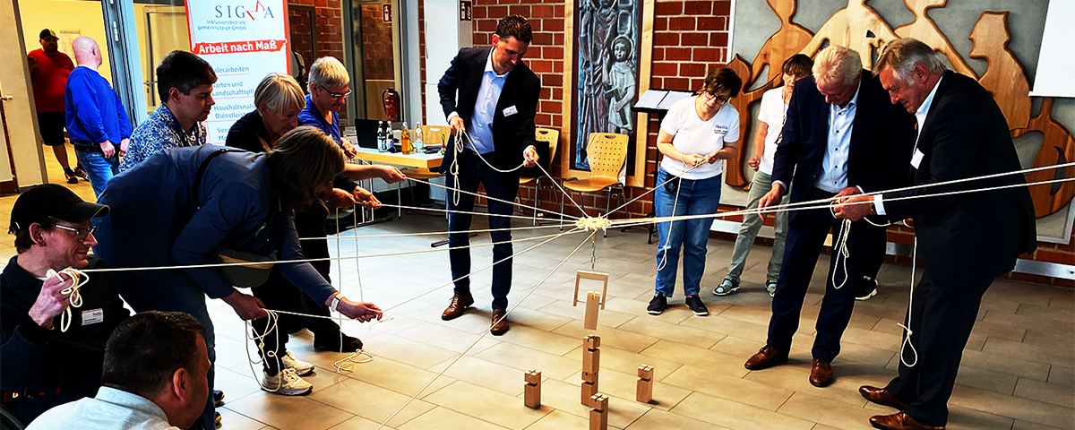 Mehrere Menschen versuchen einen Turm aus Holzblöcken zu bauen. Dazu benutzen sie einen Greifer, den man mit vielen Seilen bedient.