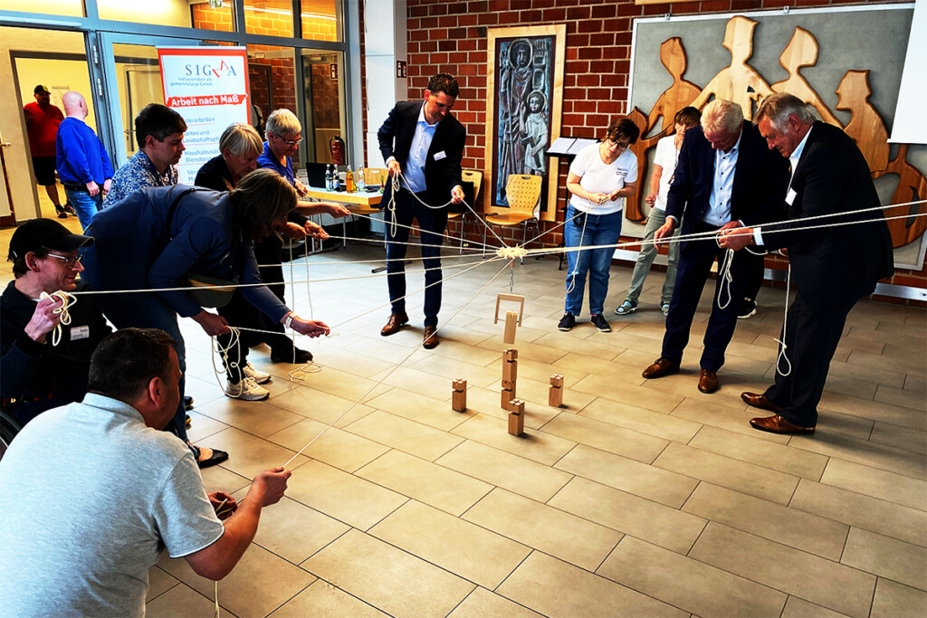 Mehrere Menschen versuchen einen Turm aus Holzblöcken zu bauen. Dazu benutzen sie einen Greifer, den man mit vielen Seilen bedient.