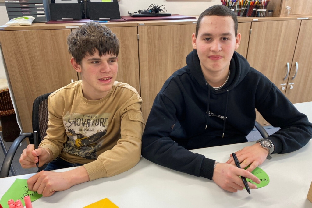 Zwei Schüler sitzen an einem Tisch und schauen in die Kamera