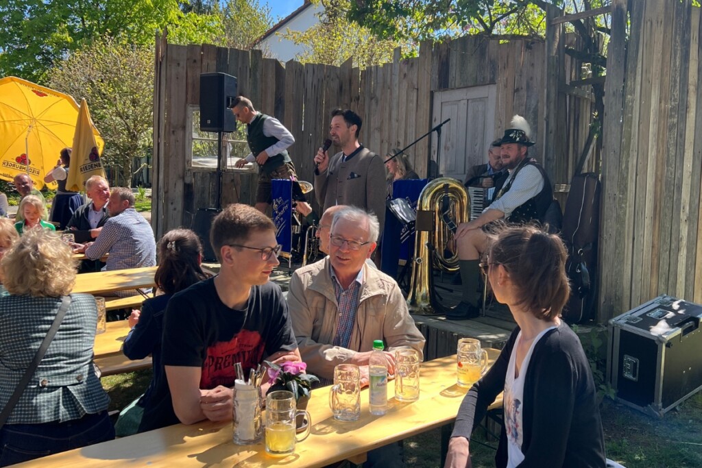 Auf dem Maibockfest am Hermannsberg spielt eine Volksmusikgruppe auf einer kleinen Bühne, während Gäste an Biertischen sitzen und Getränke genießen.
