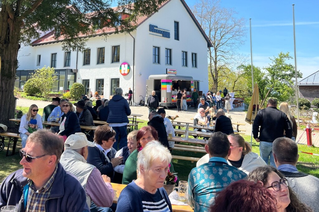Zahlreiche Besucher sitzen bei sonnigem Wetter an Biertischen vor einem Gebäude und genießen Getränke, während im Hintergrund ein Ausschankwagen und weitere Gäste zu sehen sind.