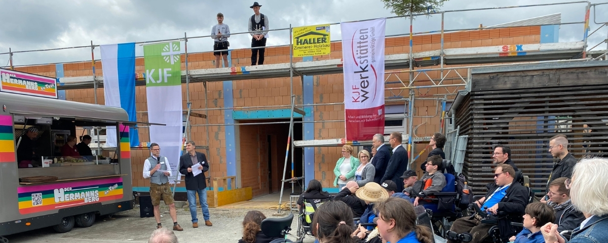 Richtfest der St. Johannes Werkstätten in Regensburg: Zwei Personen halten vor einem Neubau eine Rede, während ein großes Publikum, darunter mehrere Rollstuhlnutzende, zuhört; auf dem Gerüst hängen Fahnen und ein Richtbaum.
