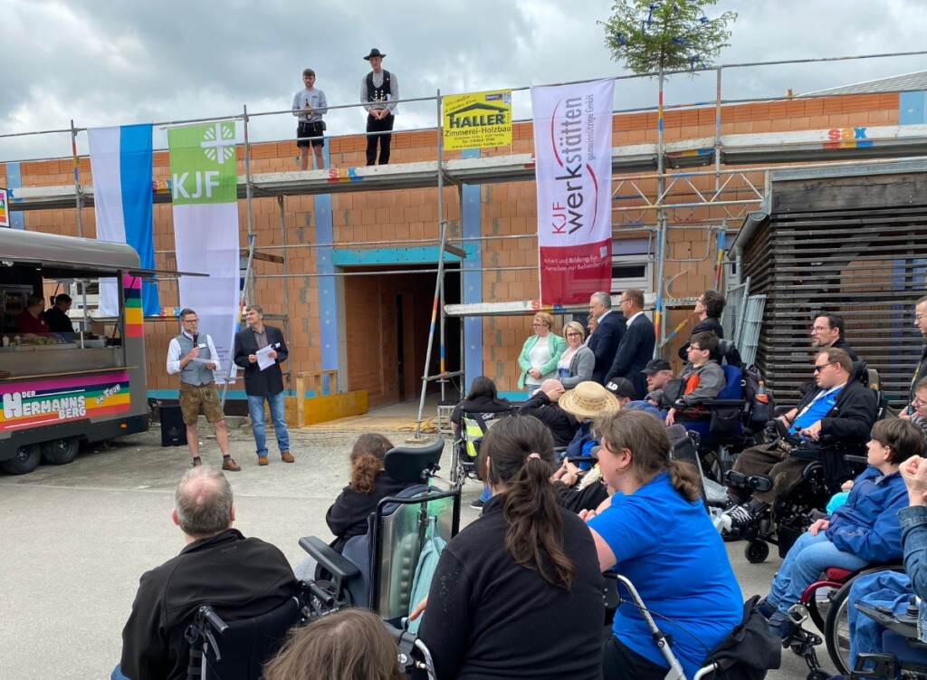 Richtfest der St. Johannes Werkstätten in Regensburg: Zwei Personen halten vor einem Neubau eine Rede, während ein großes Publikum, darunter mehrere Rollstuhlnutzende, zuhört; auf dem Gerüst hängen Fahnen und ein Richtbaum.