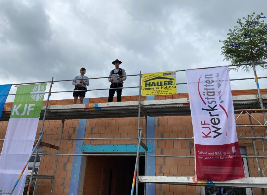 Zwei Personen stehen auf einem Baugerüst vor einem Neubau und sprechen beim Richtfest der St. Johannes Werkstätten in Regensburg den Richtfestspruch. Neben ihnen hängen Fahnen der KJF Werkstätten und ein Richtbaum ist zu sehen.