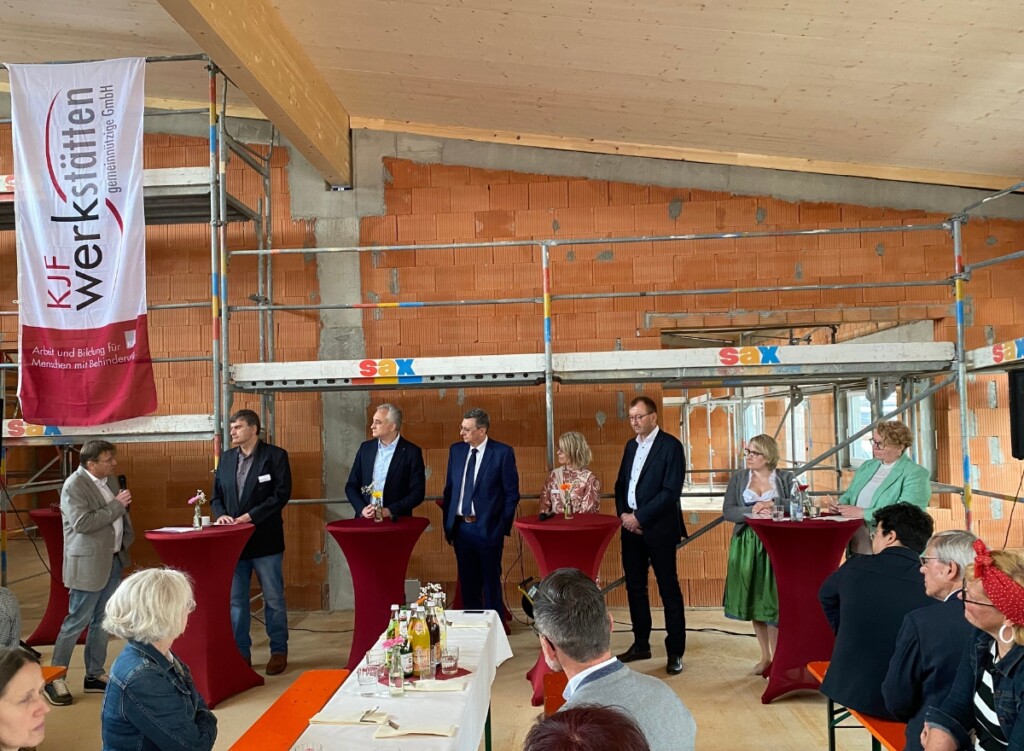 Im Innenbereich eines Rohbaus stehen mehrere Personen an Stehtischen und führen beim Richtfest der KJF Werkstätten in Regensburg eine moderierte Gesprächsrunde. Im Hintergrund sind Bauwände, ein Gerüst und eine Fahne der KJF Werkstätten zu sehen, im Vordergrund sitzt Publikum an Tischen.