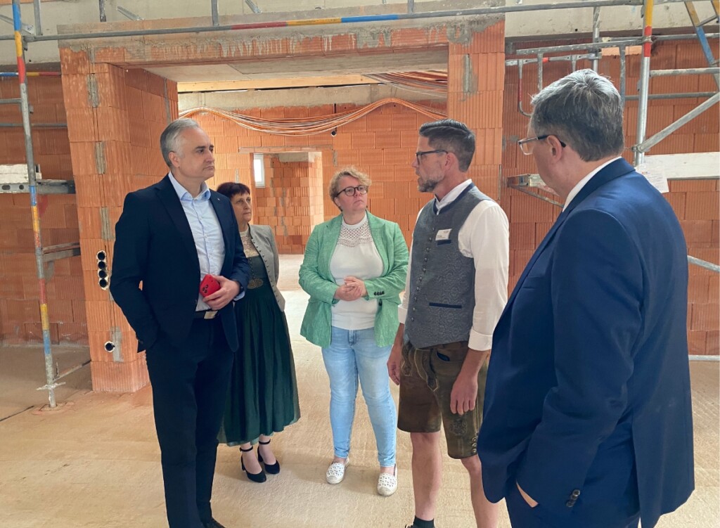 Mehrere Personen stehen im Rohbau eines Gebäudes und unterhalten sich zwischen Backsteinwänden und Baugerüst. Im Hintergrund sind offene Durchgänge, Kabel an der Decke und Baustellenstrukturen zu sehen.