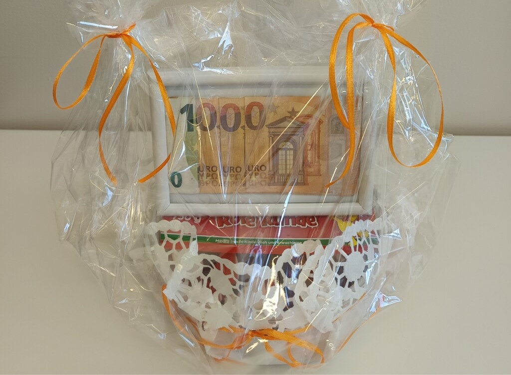 Liebevoll verpackter Geschenkkorb mit 1.000-Euro-Schein und Fruchtgummi, dekoriert mit transparentem Zellophan und orangefarbenen Schleifen als Zeichen der Wertschätzung.