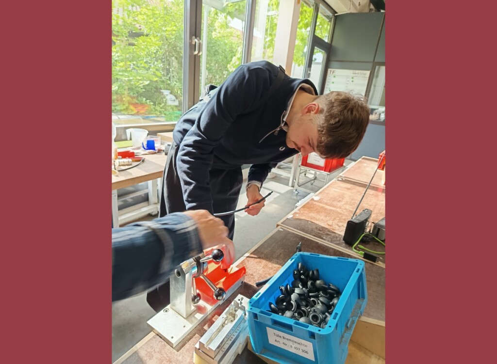 Luke Hoß arbeitet an einem Montagearbeitsplatz und setzt ein Kabel in eine Vorrichtung ein; auf dem Tisch stehen Werkzeug und eine blaue Kiste mit Bauteilen, im Hintergrund fällt Tageslicht durch große Fenster.