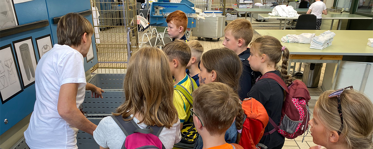 Ein Tag voller Begegnungen und neuer Perspektiven: Erika Stelzl zeigt den Schulkindern der Grundschule Haibach etwas an einem Wäschewagen; im Hintergrund Faltmaschinen, Sortiertische und gestapelte weiße Wäsche.