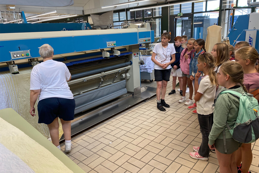 Betriebsführung in der Wäscherei: Erika Stelzl steht vor einer großen blauen Mangel und erklärt den Schulkindern die Maschine; rechts stehen neun Kinder in einer Reihe und blicken zur Maschine, links eine weitere Mitarbeiterin mit dem Rücken zur Kamera.