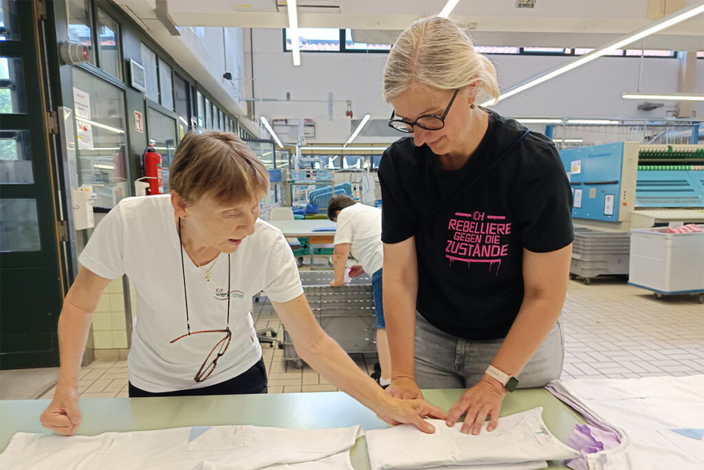 Erika Stelzl und Silke Prößl stehen an einem Tisch mit Wäsche. Erika Stelzl zeigt an einem Kleidungsstück, wie es gefaltet werden soll.