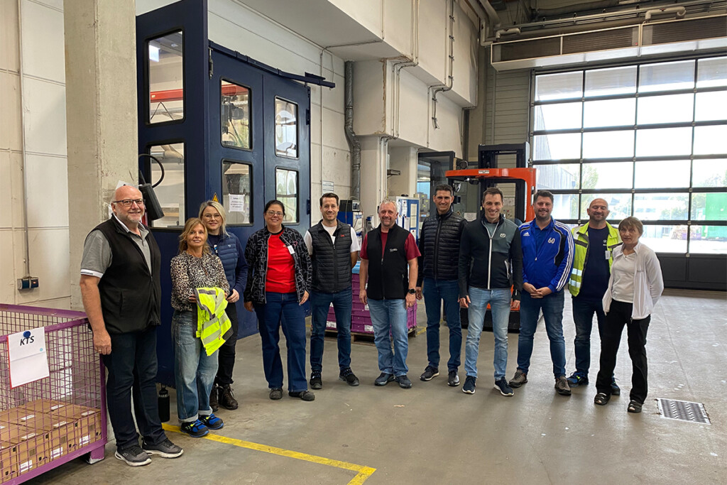 Elf Personen stehen nebeneinander für ein Gruppenfoto in einer Industriehalle. Im Hintergrund sind ein großes blaues Tor, ein orangefarbener Gabelstapler und ein breites Glas-Rolltor zu sehen.