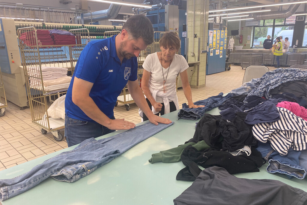 Andreas Aichinger und Erika Stelzl stehen an einem Tisch in der Wäscherei. Andreas Aichinger streicht mit beiden Händen eine Jeans glatt, während Erika Stelzl ihm dabei zuschaut. Neben ihnen liegt ein großer Haufen unsortierter Kleidung.