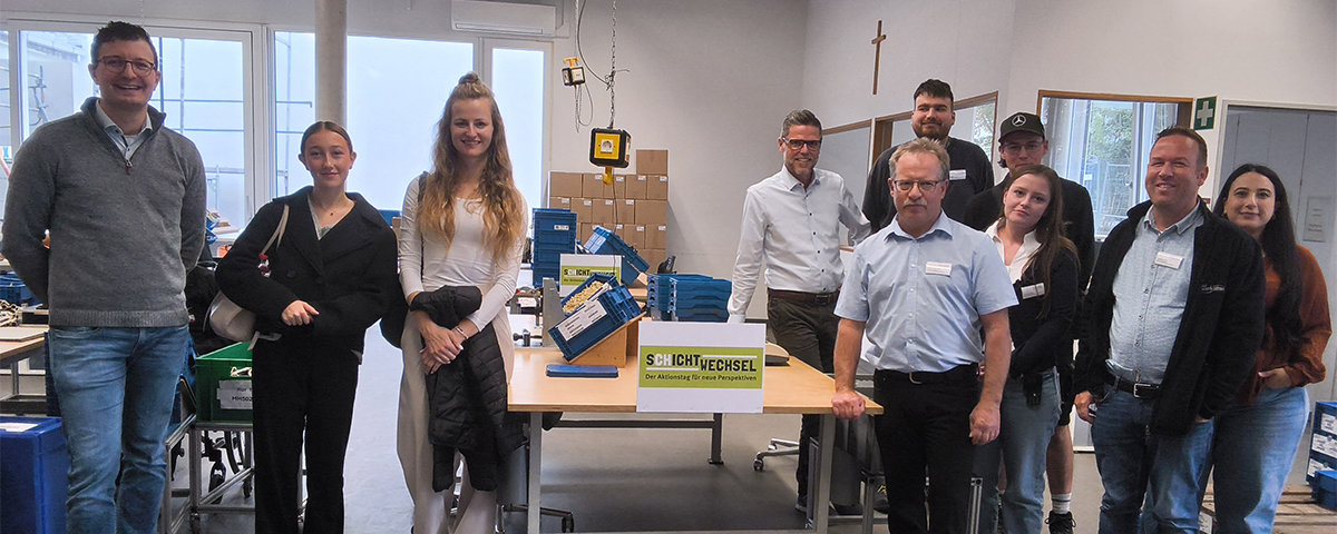 Schichtwechsel 2026 in Regensburg - Zehn Personen stehen für ein Gruppenfoto in einem hellen Arbeitsraum um einen Arbeitstisch. An der vorderen Tischkante hängt ein Schild mit der Aufschrift "SCHICHTWECHSEL - Der Aktionstag für neue Perspektiven". Die Gruppe blickt lächelnd zur Kamera, einige tragen Namensschilder.