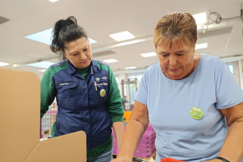 Eine Gastteilnehmerin des Aktionstags „Schichtwechsel“ arbeitet gemeinsam mit einer Beschäftigten an einem Verpackungsarbeitsplatz.