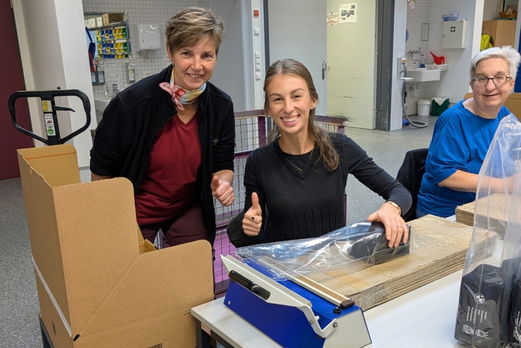 Drei Personen arbeiten gemeinsam an einem Verpackungsarbeitsplatz in der Werkstätte. Auf dem Tisch befinden sich Kartonzuschnitte, Verpackungsmaterial und ein Folienschweißgerät. Während ein Produkt verpackt wird, stehen weitere Kartons und fertig vorbereitete Materialien für den Versand bereit. Im Hintergrund sind weitere Arbeitsbereiche der Werkstätte zu sehen.