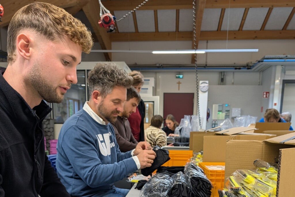 Mehrere Gäste arbeiten gemeinsam an einem Verpackungsarbeitsplatz in der Werkstätte. Auf dem Tisch befinden sich Kartons, Kunststoffverpackungen und weitere Materialien für die Konfektionierung. Im Hintergrund sind weitere Beschäftigte und Arbeitsbereiche der Werkstätte zu sehen.
