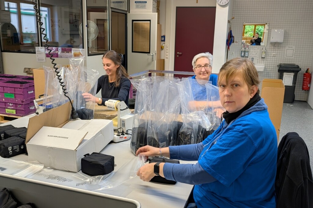 Mehrere Beschäftigte arbeiten an einem Verpackungsarbeitsplatz in der Werkstätte. Auf dem Tisch stehen Kartons, Verpackungsmaterial und bereits verpackte Produkte in transparenten Beuteln. Die Beschäftigten verpacken und bereiten die Produkte für den Versand vor. Im Hintergrund sind weitere Arbeitsbereiche der Werkstätte zu sehen.