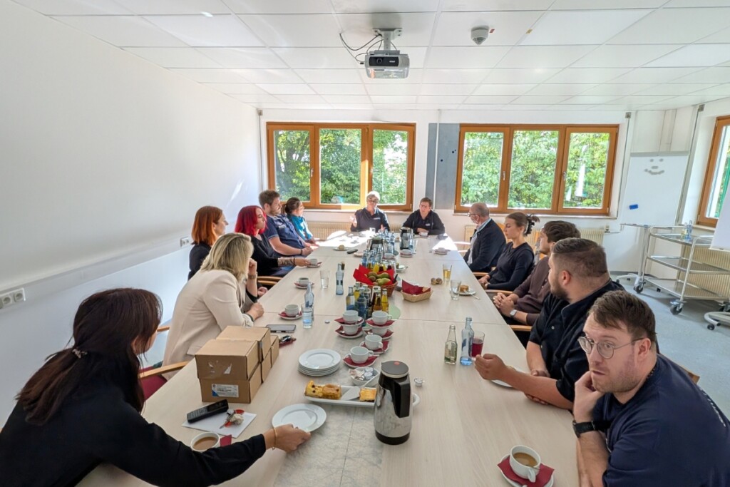 Beschäftigte und Gäste sitzen gemeinsam an einem großen Tisch in einem Besprechungsraum der Werkstätte. Die Gruppe befindet sich im gemeinsamen Austausch und Gespräch. Auf dem Tisch stehen Getränke, Kaffeegeschirr und kleine Snacks. Große Fenster sorgen für eine helle und offene Atmosphäre.