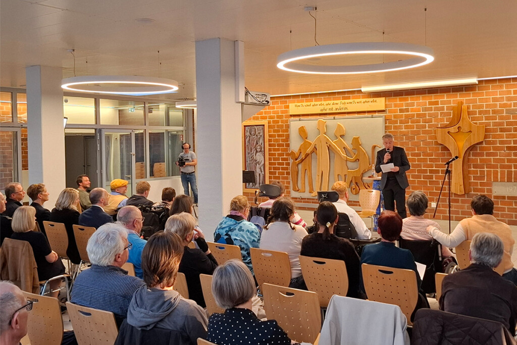 Prof. Dr. Christian Zürner steht im dunklen Anzug vor einer Backsteinwand und spricht in ein Mikrofon zu einem sitzenden Publikum, darunter auch Personen im Rollstuhl. An der Wand hängen ein Holzrelief stilisierter Menschen und ein Schild mit dem Spruch 'Wenn Du schnell gehen willst, gehe alleine. Doch wenn Du weit gehen willst, gehe mit anderen.' Im Hintergrund filmt ein Mann mit Kamera.