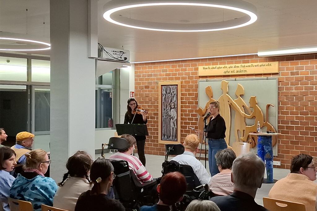 Maike Berndt-Zürner spielt Violine, während Henriette Heine in ein Mikrofon spricht. Das Publikum ist aufmerksam: Alle blicken nach vorn, zwei Personen sitzen im Rollstuhl. Im Hintergrund hängen ein Holzrelief stilisierter Menschen und ein Schild mit dem Spruch 'Wenn Du schnell gehen willst, gehe alleine. Doch wenn Du weit gehen willst, gehe mit anderen.'