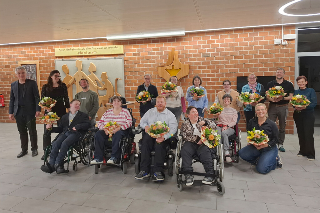 Gruppenfoto von 16 Personen in einem Saal vor einer Backsteinwand. Fünf Personen sitzen im Rollstuhl, eine kniet, zehn stehen dahinter. 14 von ihnen halten einen Blumenstrauß. Die Gruppe wirkt fröhlich: fast alle blicken lächelnd in die Kamera. An der Wand steht auf einem Schild: "Wenn Du schnell gehen willst, gehe alleine. Doch wenn Du weit gehen willst, gehe mit anderen."