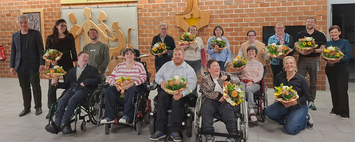 Inklusive Gedichtlesung - Gruppenfoto von 16 Personen in einem Saal vor einer Backsteinwand. Fünf Personen sitzen im Rollstuhl, eine kniet, zehn stehen dahinter. 14 von ihnen halten einen Blumenstrauß. Die Gruppe wirkt fröhlich: fast alle blicken lächelnd in die Kamera. An der Wand steht auf einem Schild: "Wenn Du schnell gehen willst, gehe alleine. Doch wenn Du weit gehen willst, gehe mit anderen."